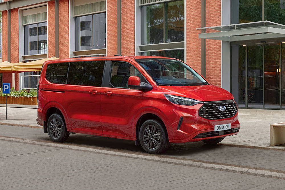 Un Ford Tourneo Custom roșu este parcat pe o stradă din oraș în fața unei clădiri din cărămidă