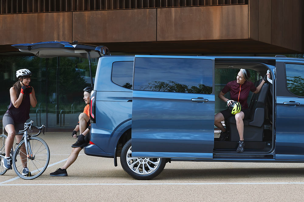 Ford Tourneo Custom albastru parcat cu ușile laterale deschise și câteva persoane cu biciclete în apropiere.