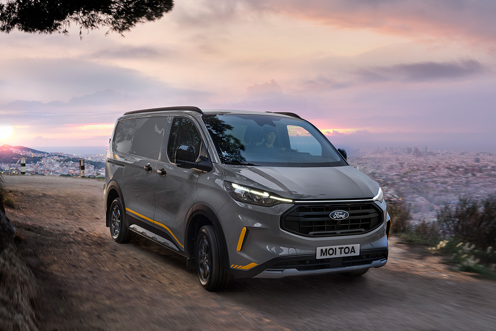 Grey Ford Transit Custom Trail van with yellow accents drives on rough road.