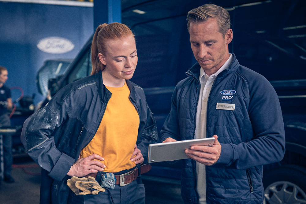 Doi tehnicieni într-un garaj care se uită la o tabletă în fața unui furgon Ford E-Transit.