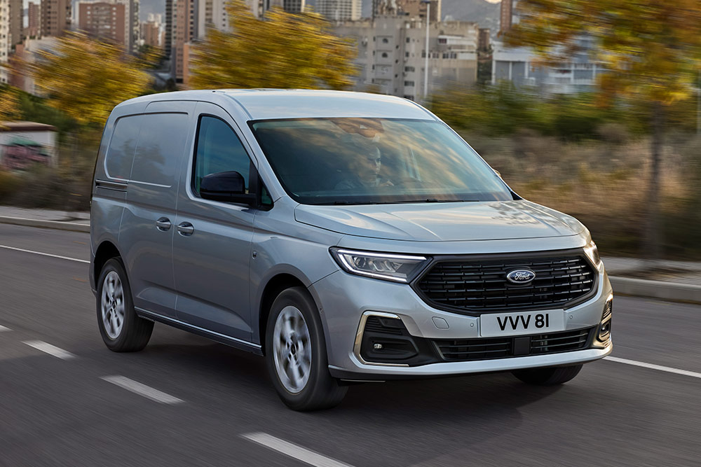 Un Ford Transit Connect deplasându-se într-un oraș, prezentând ramele cromate ale proiectoarelor de ceață și farurile automate.