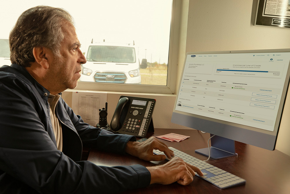 O persoană așezată la birou analizează date pe un monitor, cu o dubă Ford vizibilă prin fereastră.