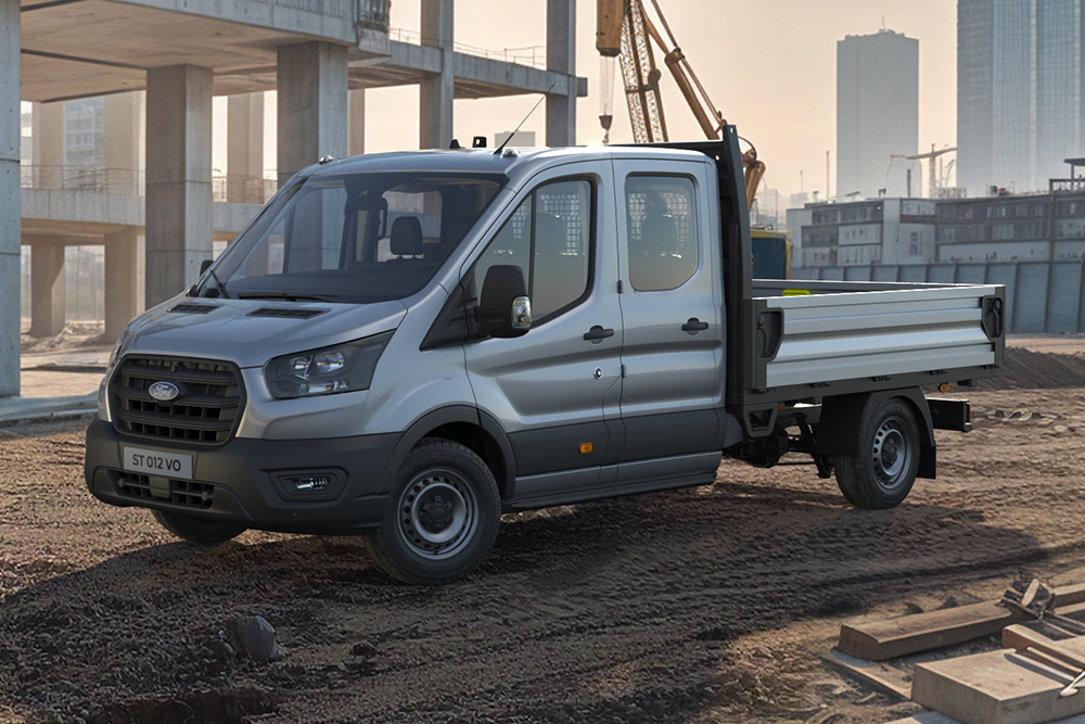 Un Ford Transit basculantă cu cabină dublă, argintiu, staționează pe un șantier, cu o macara în fundal.