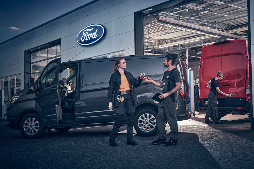Doi angajați Ford își predau cheile unei dube, cu o altă dubă roșie în service, la lăsarea serii.